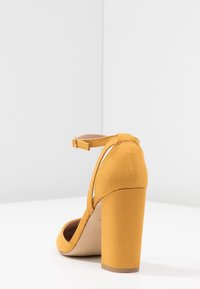 Musztardowe żółte szpilki wykonane z zamszu, z grubym obcasem w kształcie bloku, paskiem wokół kostki z klamrą oraz otwartym noskiem.