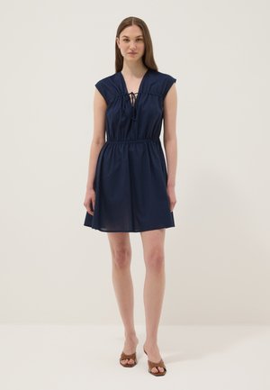 Mujer de pie con un vestido azul marino sin mangas, con cintura fruncida y detalle de lazo delantero, combinado con tacones abiertos marrones.