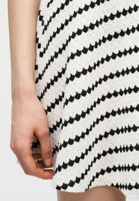 Χέρι που ακουμπά δίπλα σε λευκή υφασμάτινη φούστα με διαγώνιες μαύρες ζιγκ-ζαγκ ρίγες.