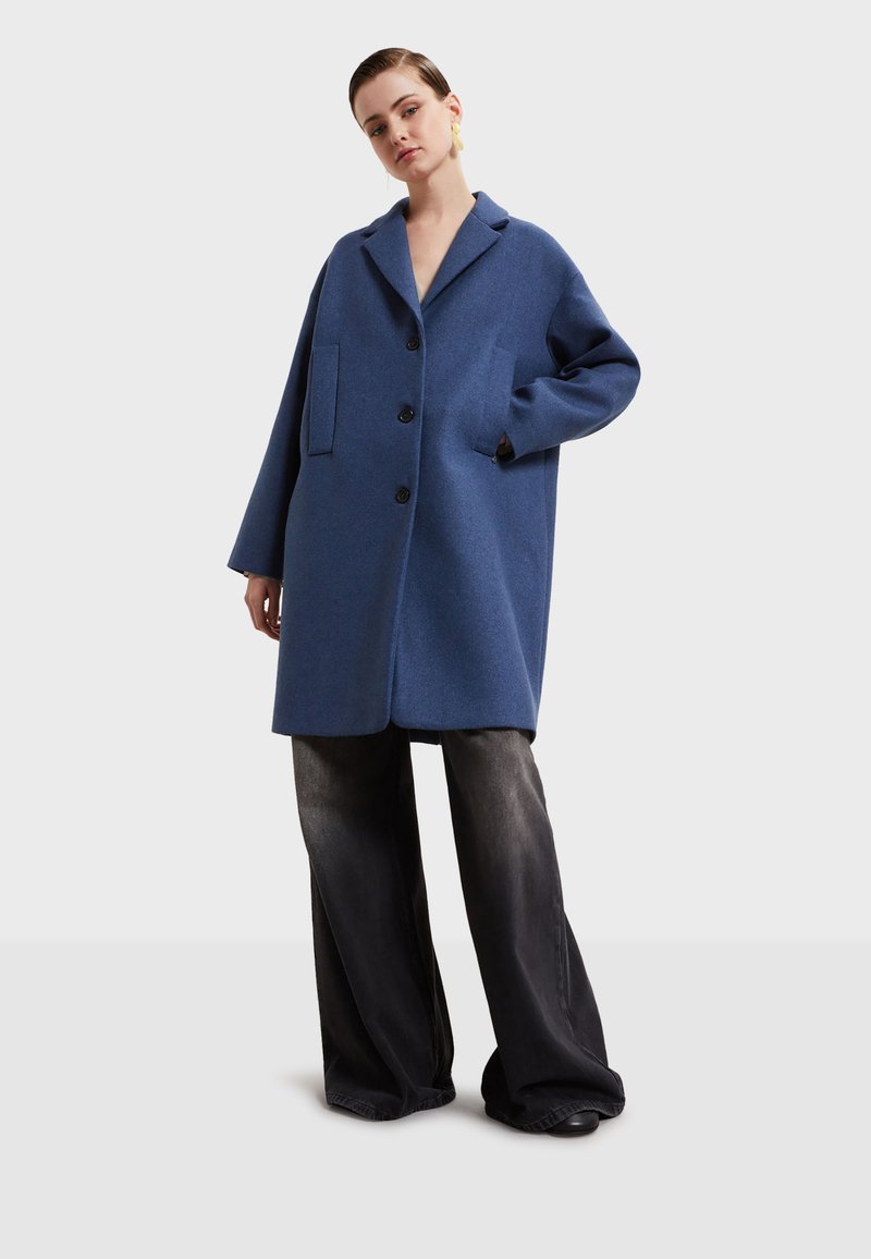 Modrý vlnený kabát s klopovým golierom, zapínaním na gombík a dvoma prednými vreckami. Má voľný strih a dlhé rukávy.