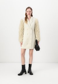 Manteau double boutonnage crème avec des manches texturées et duveteuses et de grands boutons, associé à des bottines noires et un petit sac à main noir.