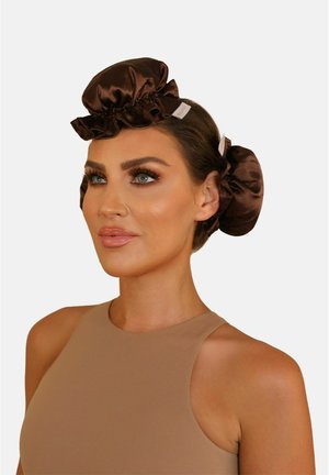 Vrouw met bruine satijnen hairbonnets, gestyled bovenop en aan de zijkant van haar hoofd, gekleed in een beige mouwloze top, met een natuurlijke make-up.