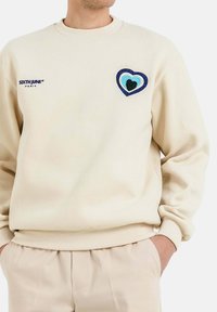 Sudadera beige con un diseño de corazón bordado en azul, verde azulado y verde. Cuello y puños de canalé; logo de la marca en el pecho.
