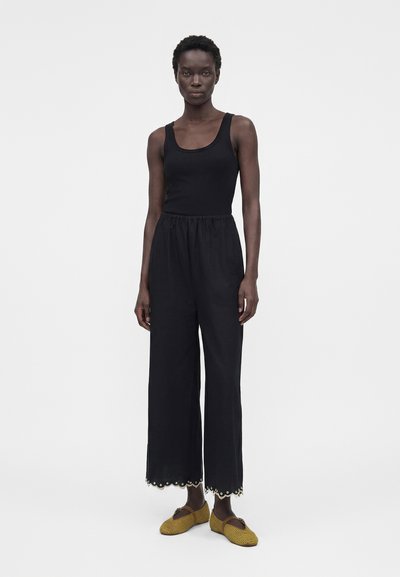 Γυναίκα στέκεται φορώντας μαύρο αμάνικο τοπ και φαρδιά μαύρη παντελόνα με λεπτομέρειες από κορδέλα με τρύπες, συνδυασμένη με πλεκτά φλατ παπούτσια σε μουσταρδί κίτρινο.