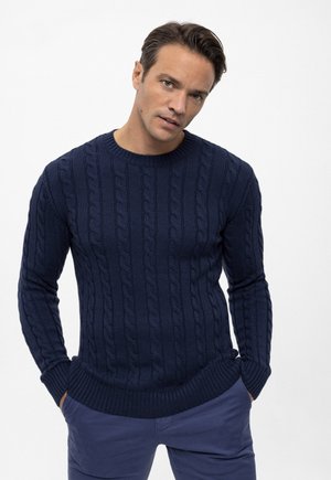 Marineblauer, kabelstrick gestrickter Pullover mit rundem Ausschnitt und gerippten Bündchen und Saum, getragen mit maßgeschneiderten blauen Hosen. Strukturmuster über das gesamte Kleidungsstück.