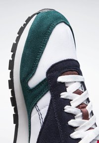 Sneaker med en hvit mesh-overdel med mørk marineblå og grønn semsket skinn akenter, teksturert gummisåle, og kontrasterende snøringer i kobber.