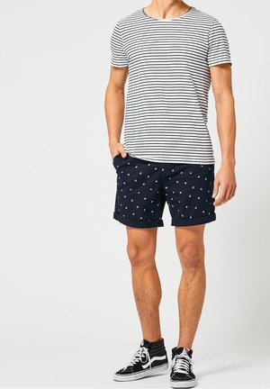 Man draagt een wit-zwart gestreept T-shirt, marineblauwe korte broek met patroon en zwarte hoge sneakers met witte veters, staat met één hand in de zak.