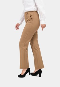 Lichtbruine, lichte broek met een elastische tailleband, zijzakken, rechte pijpen en subtiele stikseldetails, gecombineerd met zwarte hoge hakken.