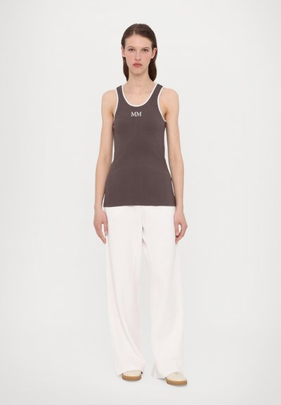 Brūns ribots topiņš ar baltu apmalīti un "MM" logo, kombinēts ar brīviem baltiem biksēm un gaismas krāsas apaviem.