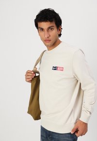 Beige genser med rund hals laget av bomull, med en logo i rødt, hvitt og blått. Modellen holder en brun totebag.
