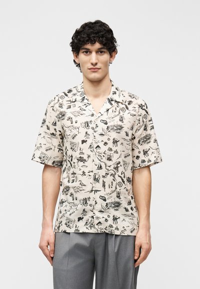 Jeune homme aux cheveux bouclés portant une chemise beige à manches courtes avec des croquis noirs sur le thème du voyage et un pantalon gris, debout devant un fond neutre.