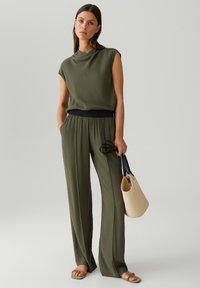 Olijfgroene, losvallende top met een kraaghals, gecombineerd met bijpassende wijde broek. Het model houdt een zwarte schoudertas vast en draagt beige sandalen.