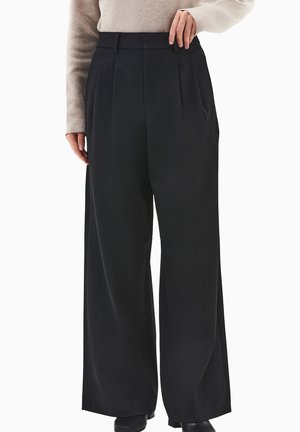 Czarne spodnie o szerokich nogawkach z wyraźnymi plisami, wysokim stanem i gładką fakturą tkaniny. Stylizowane z jasnobarwną bluzką z długim rękawem.