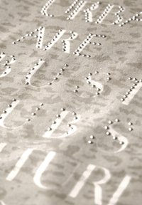 Tejido con un fondo beige claro que presenta tachuelas plateadas elevadas formando letras. Textura suave con un sutil patrón bajo el texto.