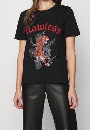 Femme portant un t-shirt noir avec texte rouge « Flawless » et un motif de tigre rugissant sur un design floral blanc, associé à un pantalon en cuir noir.