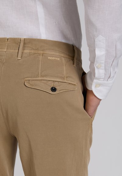 Close-up van een persoon die beige Incotex-broek draagt met een achterzak met knoop en een wit overhemd met lange mouwen, hand in de voorzak.
