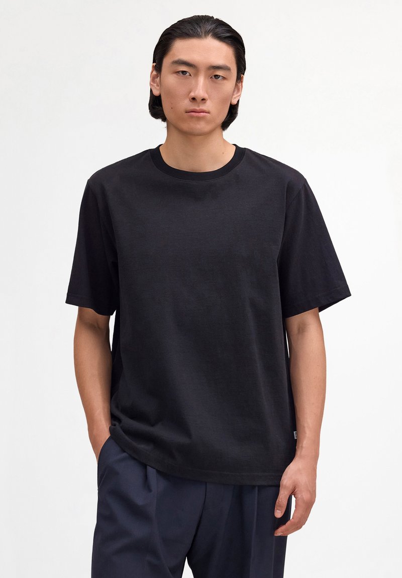 Junger Mann mit schwarzen Haaren, der ein lockeres schwarzes T-Shirt und dunkle Hosen trägt, vor einem schlichten weißen Hintergrund stehend.