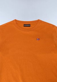 T-shirt en coton orange à col rond, avec un petit logo brodé du drapeau norvégien sur le côté gauche de la poitrine. Texture lisse et design simple.