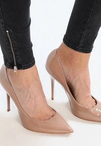 Tacones altos de charol en tono nude con puntas puntiagudas y un diseño elegante, con tacones de aguja y un acabado brillante, combinados con jeans negros.