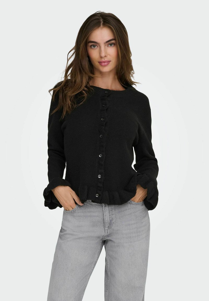 Pull noir à boutons avec un col rond, des poignets et un ourlet à volants. Fabriqué en tissu texturé. Associé à un jean gris clair.