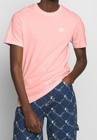 T-shirt in cotone rosa con scollatura a girocollo. Presenta un logo Nike bianco sul petto sinistro. Indossato con pantaloni shorts in denim a fantasia.