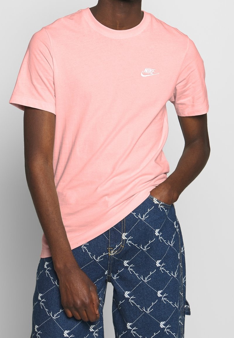 T-shirt in cotone rosa con scollatura a girocollo. Presenta un logo Nike bianco sul petto sinistro. Indossato con pantaloni shorts in denim a fantasia.