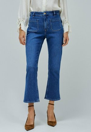 Jeans in denim blu a zampa d'elefante con vita alta, tasche frontali evidenti e orlo leggermente sfilacciato, abbinati a scarpe con tacco alto marroni.