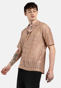 Camicia a maniche corte in tessuto perforato rosa chiaro con motivi a diamante, dotata di colletto e bottoni, indossata con pantaloni neri.