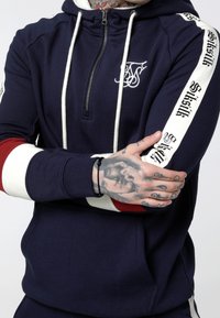 Sudadera con cremallera en color navy, con cordones blancos, que presenta un logo negro y rayas en contraste en rojo y blanco en las mangas. Tejido suave y texturizado.