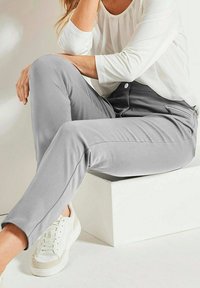Grijze broek met een slank ontwerp en een zijzak met rits, gedragen met een witte lange mouw top en witte sneakers. Zachte stoftextuur.