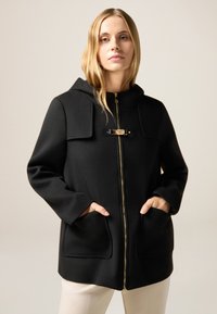 Abrigo negro con capucha y cierre frontal de cremallera, con dos bolsillos laterales, detalles en dorado y tejido texturizado. Líneas limpias y diseño minimalista.