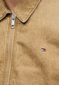 Barna, texturált anyagból készült, zipzáras, galléros dzseki. A mellkasán kis, kék, fehér és piros hímzett logó látható.