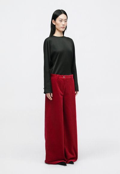 Black long-sleeve top paired with wide-leg red corduroy pants featuring textured stripes. Model stands in front of a plain background.