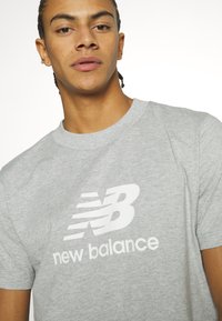 Pilkos medvilnės marškinėliai su ryškiu baltu "NB" logotipu ir "new balance" tekstu po juo. Apvalus iškirpimas ir trumpomis rankovėmis.