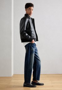 Schwarze Lederjacke mit Fronttaschen, darunter ein weißes Hemd, lockere blaue Jeans und schwarze Slipper, präsentiert vor einem neutralen Hintergrund.