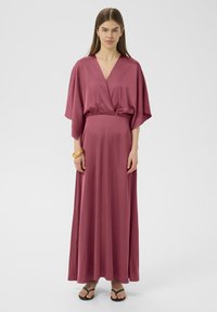 Fließendes, tiefrosa Kleid mit V-Ausschnitt, Fledermausärmeln und taillierter Taille, aus glattem Stoff. Getragen mit schwarzen Flip-Flops.