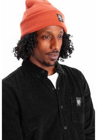 Rust-colored ribbed beanie with a black logo patch, worn over a black corduroy shirt featuring a pocket and a button-up design.