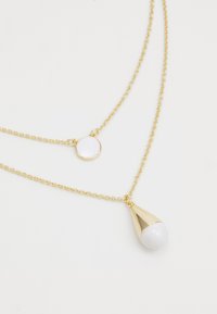 Collana a più strati in tono oro con un pendente rotondo bianco e un pendente a goccia bianco. Le catene hanno un design a maglie delicate.