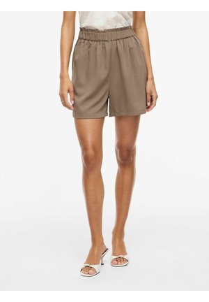 Femme portant un short marron ample à taille élastique associé à des sandales blanches à talons et bouts ouverts sur un fond uni.