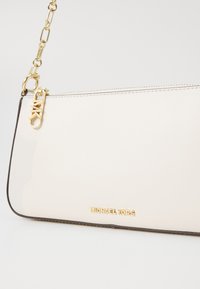 Beige synthetic handbag with gold hardware, zip closure, and embossed logo. Features a chain strap and smooth texture with a contrasting edge.