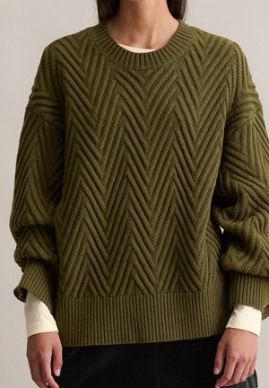Personne portant un pull en maille épaisse vert olive avec un motif chevron par-dessus une chemise blanche à manches longues, les mains détendues le long du corps.