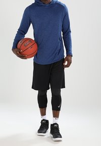 Maglietta sportiva a maniche lunghe blu con cappuccio, pantaloni corti neri in rete, scarpe da ginnastica nere Nike, ginocchiere nere, mentre tiene un pallone da basket.