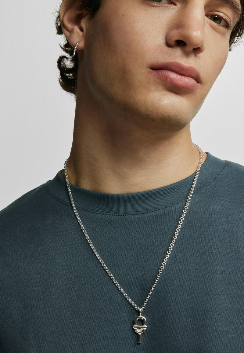 Silberne Halskette mit einem ovalen Anhänger, der ein Schlüsselmotiv zeigt, getragen über einem dunkeltealfarbenen Rundhalsausschnitt-Shirt.