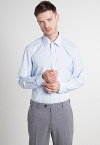 Svijetloplava košulja dugih rukava s gumbima i oštrim kragnom, u kombinaciji s sivim hlačama s uzorkom. Košulja ima glatku teksturu i ravnu kroj.