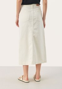 Jupe en denim crème, longueur genou, avec une coupe droite, deux poches arrière et une couture centrale à l'arrière. Associée à des sandales plates.