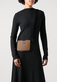 Hnedá textúrovaná crossbody taška s čiernym popruhom a zipsovým uzáverom. Obdĺžnikový tvar, s kovovým logom na prednej strane.