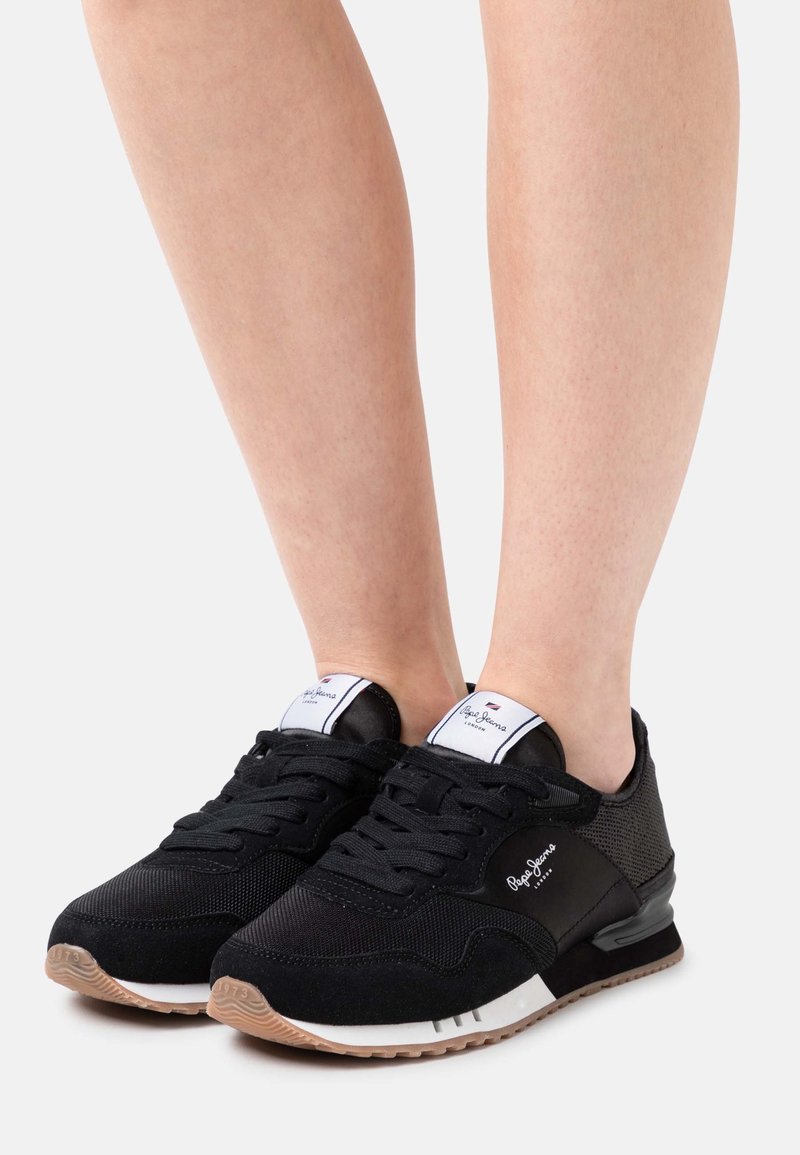 Black Pepe Jeans sneakers with white branded tongue and beige soles worn on bare feet against white background.