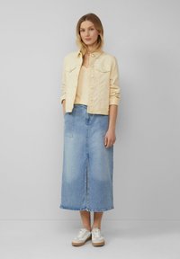 Lichtgele denimjacket over een beige top, gecombineerd met een lange blauwe denimrok en witte sneakers met lichtbruine zolen.
