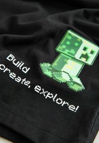 Čierne bavlnené tričko s pixelovanou zelenou postavičkou a bielym textom: „Stavaj, tvor, objavuj!“ Text je blokový a postavička detailná.