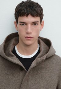 Cappotto di lana marrone con un ampio cappuccio e chiusura con bottoni, indossato sopra un maglione nero e una t-shirt bianca, caratterizzato da una texture morbida.